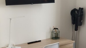 A workspace with a wooden desk, a white chair, a flat-screen TV mounted on the wall, a small decorative item on the desk, and a vacuum cleaner against the wall.