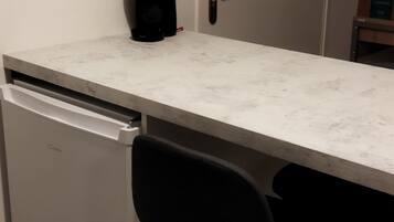 Une cuisine de taille réduite avec un plan de travail blanc, une chaise noire et un réfrigérateur blanc.