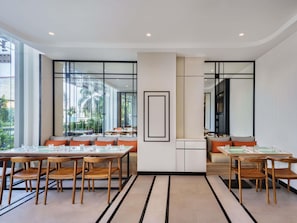 Un comedor moderno con mesas y sillas de madera, ventanas amplias con vistas a palmeras y un pilar blanco central con un cuadro enmarcado.