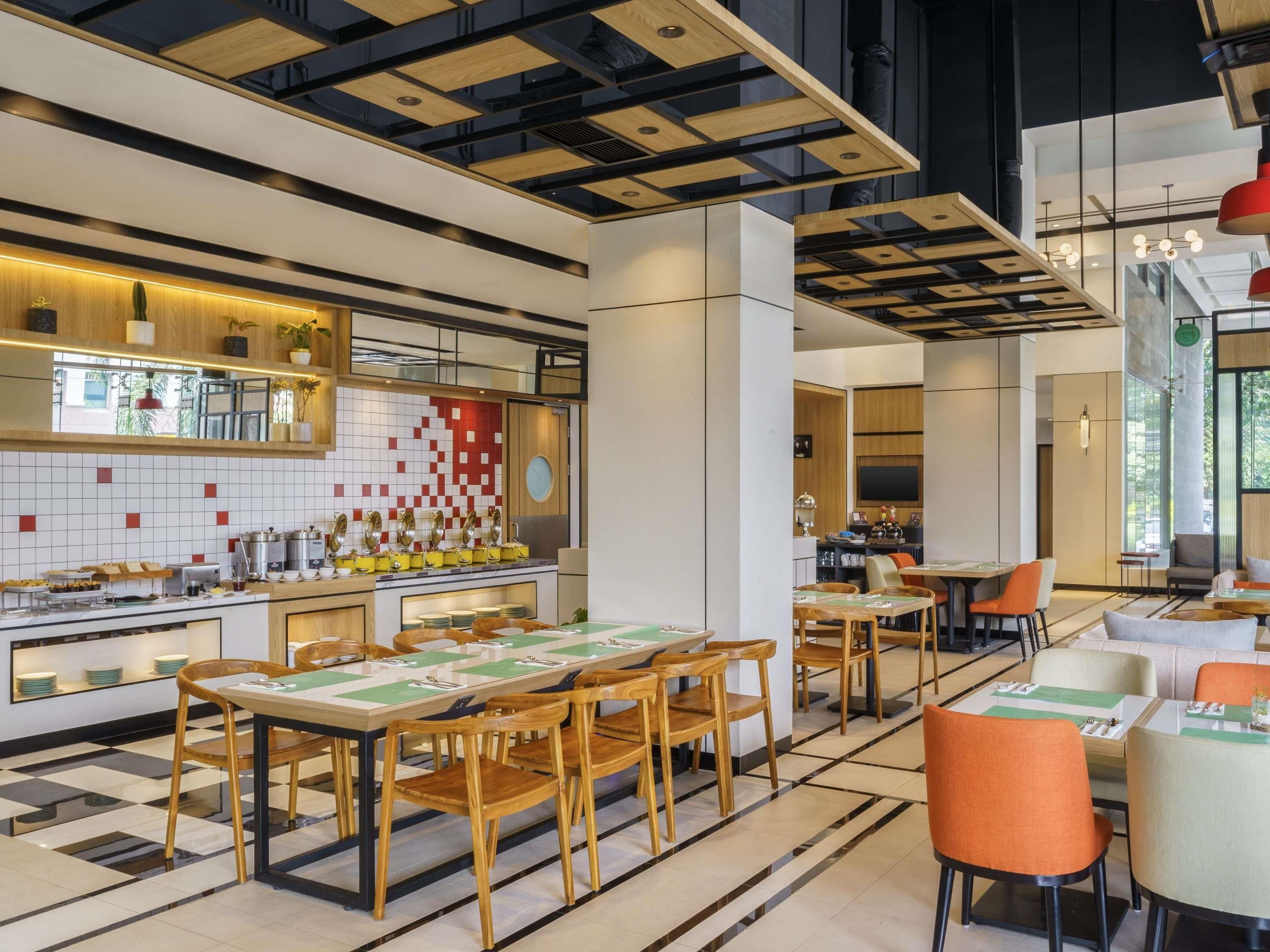 A modern restaurant interior with a long communal dining table, individual tables, and a kitchen area with a tiled backsplash.