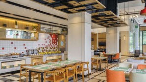 Een modern restaurantinterieur met een lange gedeelde eettafel, losse tafels en een keukenhoek met een betegelde muur.