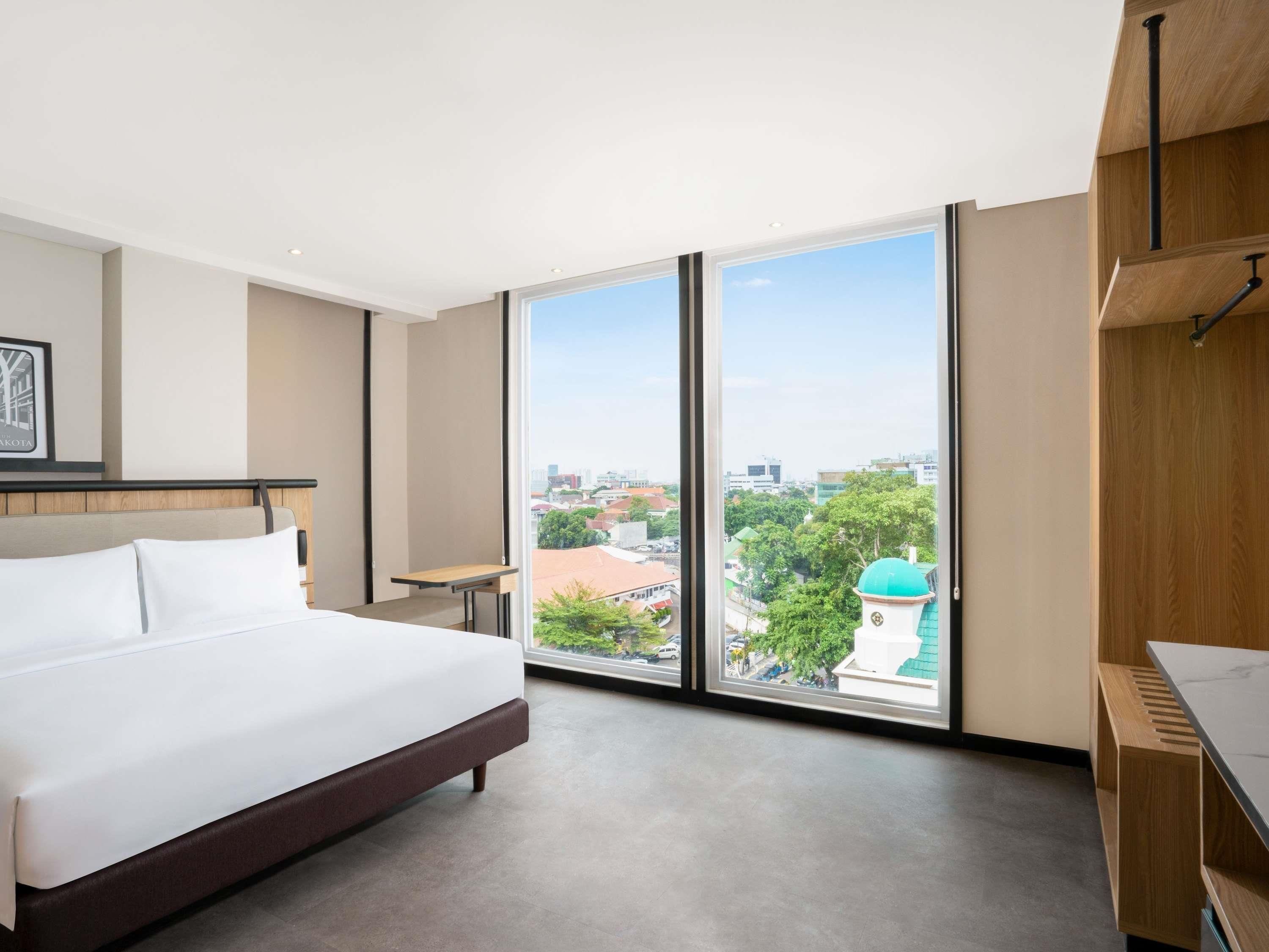 A hotel room with a large bed, a desk, and a view of the city through floor-to-ceiling windows.