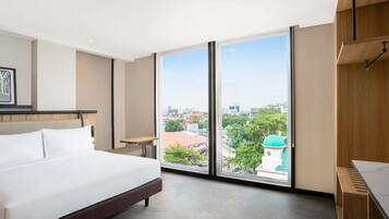 A hotel room with a large bed, a desk, and a view of the city through floor-to-ceiling windows.