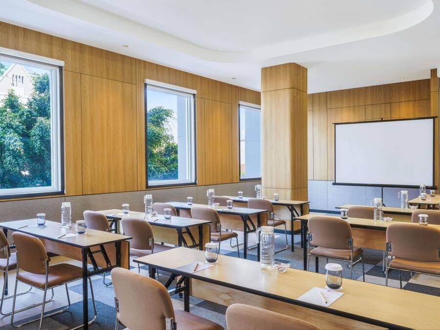 A conference room with tables, chairs, a projector screen, and large windows with a view of trees.