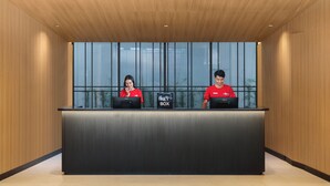 Un mostrador de recepción con dos empleados uniformados de rojo, un buzón de llaves y vistas a zonas verdes a través de amplios ventanales.