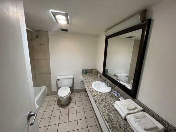 A bathroom with a toilet, sink, mirror, and a shower area. - Boardwalk Suites Lafayette (Lafayette)