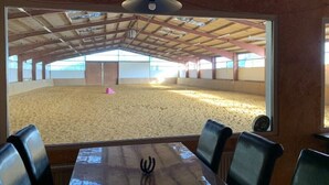 Une carrière d’équitation intérieure avec un sol sablonneux, des poutres et une structure de plafond apparents, et une table avec des chaises au premier plan.