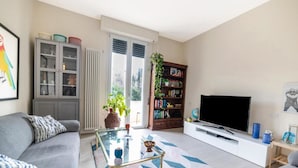 A living room with a sofa, a coffee table, a bookshelf, a TV, and a window with blinds. - Tommaso Apartment with free parking (Firenze)