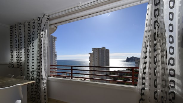 A room with a balcony overlooking a coastal view, featuring a dining table and chairs.