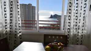 A dining area with a table and a basket of fruit, a window with a view of the sea and buildings, and curtains with a pattern.