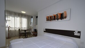 A hotel room with a large bed, a desk, and a chair. There is a painting on the wall and a window with curtains.