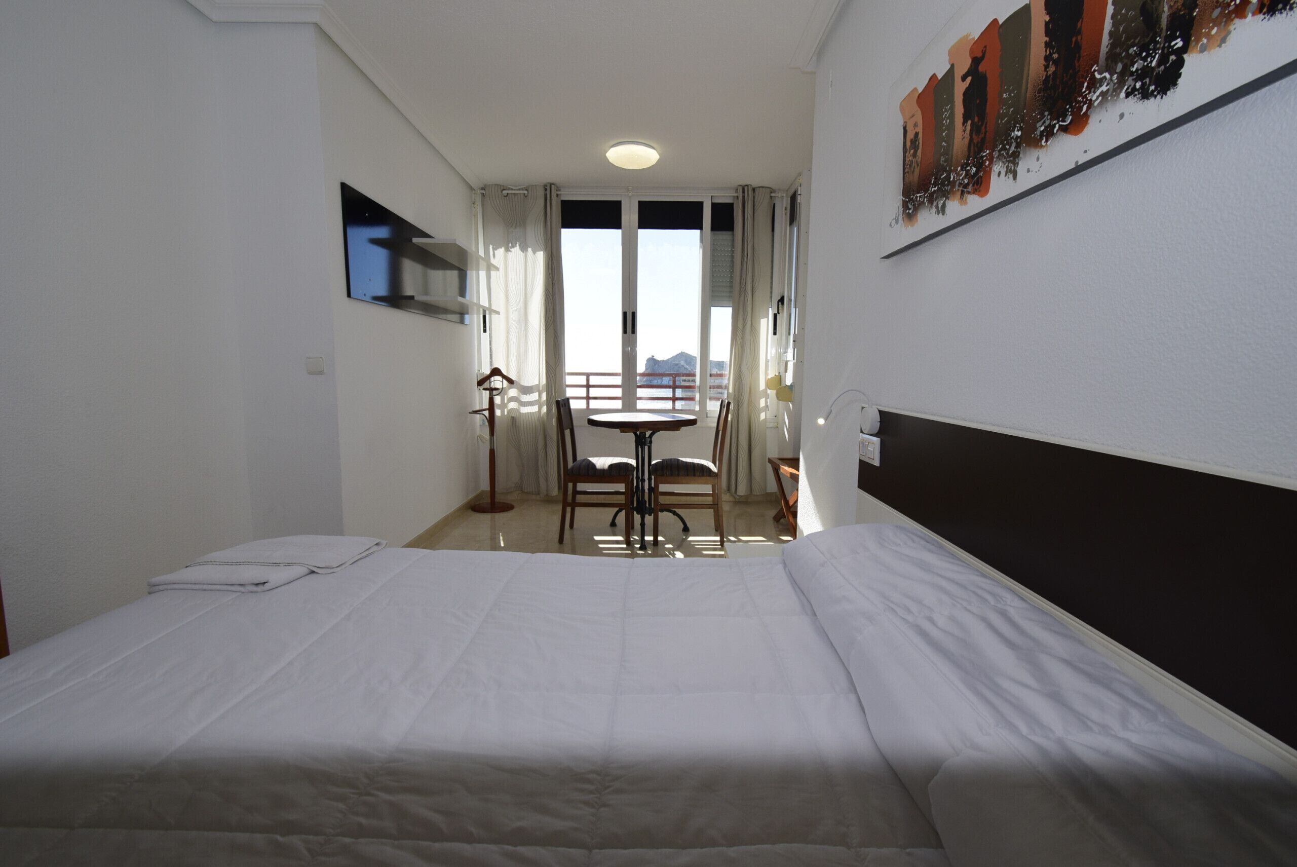 A hotel room with a bed, a small table with chairs, a view of the outdoors, and a painting on the wall.