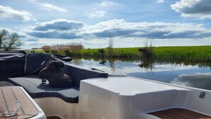 Cubierta de barco con una mesa y zona cubierta, rodeada de agua tranquila y un campo verde bajo un cielo nublado.