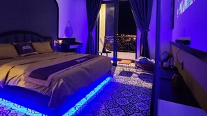 A modern bedroom with a bed, a nightstand, a wall-mounted projector, and a balcony with a railing. - Punita Hotel (Da Nang)