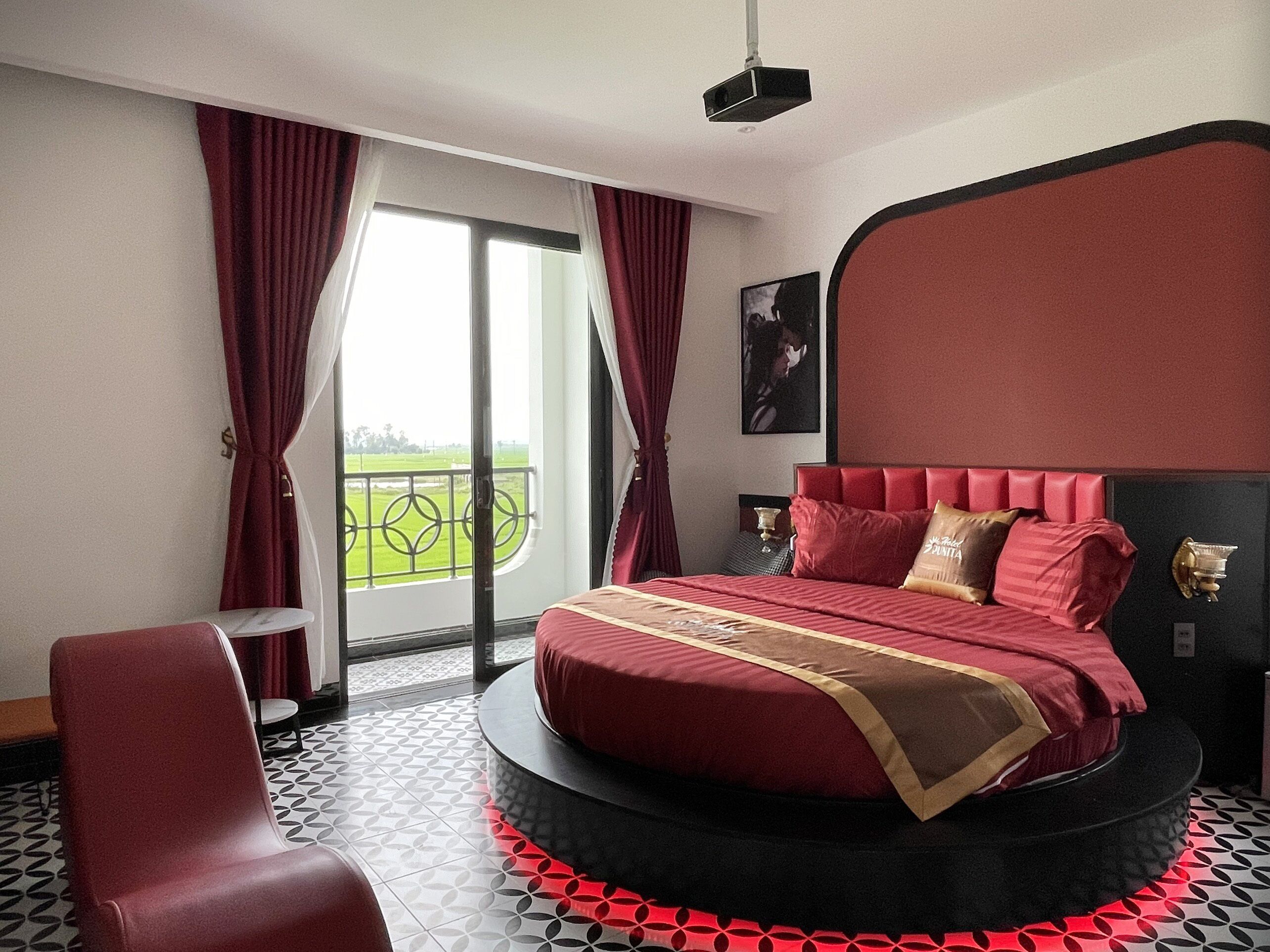 A bedroom with a circular bed, red curtains, a view of a green landscape, and a patterned floor.