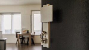 A door with a keyhole and a padlock, a dining area with a table and chairs, and a window allowing natural light. - Appartement 18 wemeldinge (Wemeldinge)