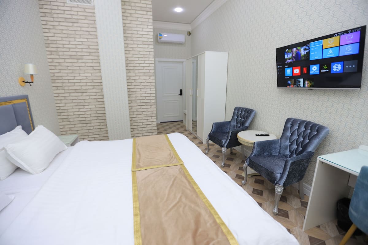 A hotel room with a bed, two armchairs, a small table, and a TV mounted on the wall displaying various apps.