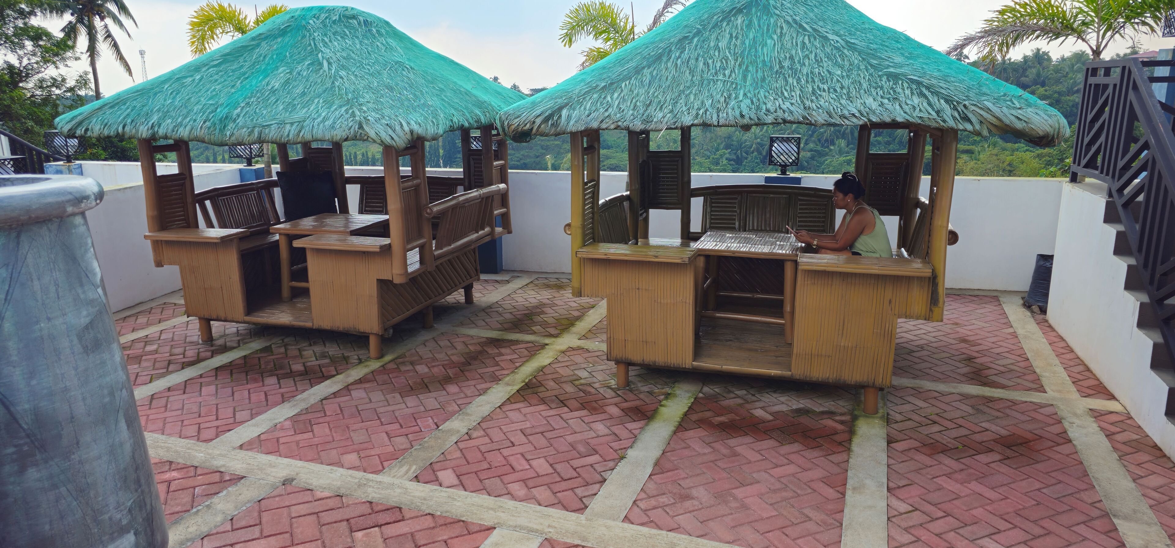 Two thatched-roof wooden structures with seating, a tiled floor, and a view of greenery.
