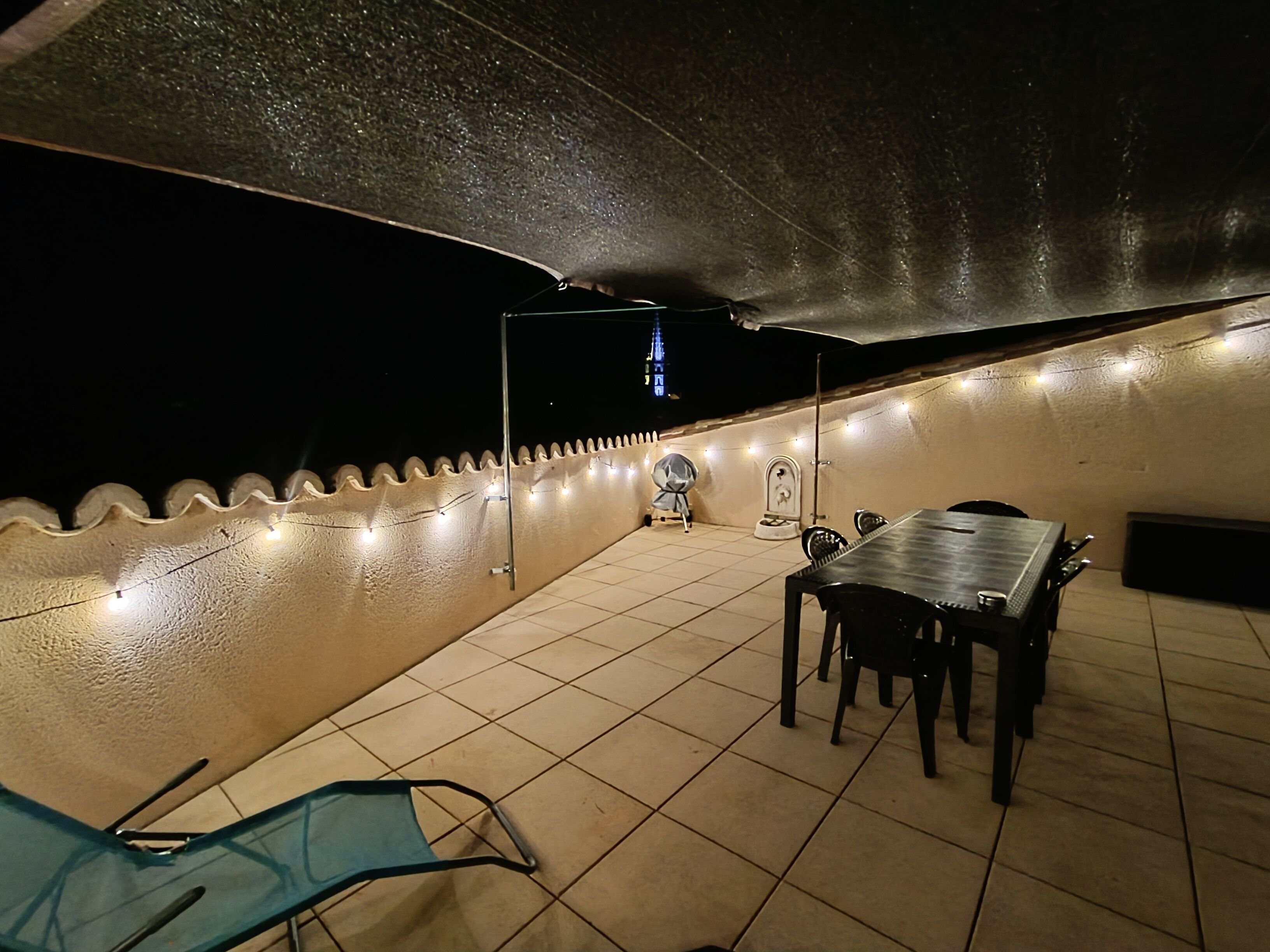 Een terras buiten met betegelde vloer, een tafel en stoelen, een overdekte zithoek en lichtslingers.
