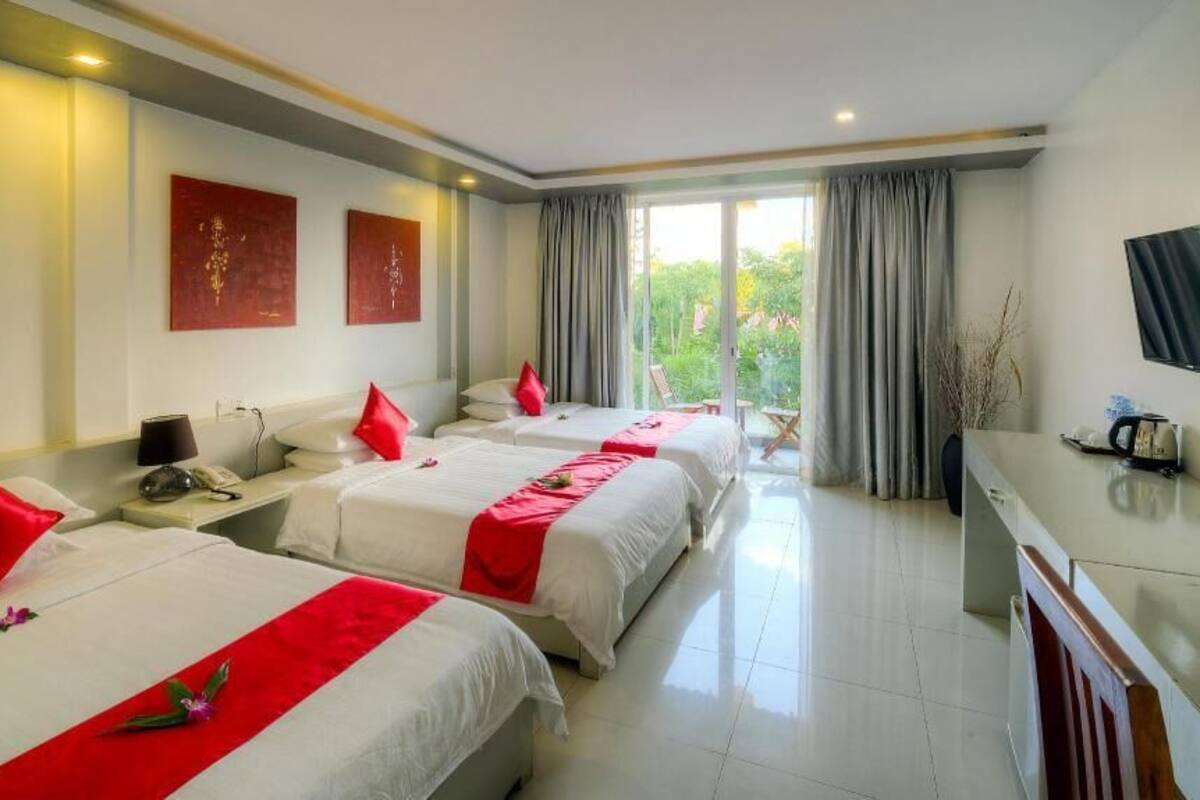 A hotel room with three beds, a desk, a television, and a view of greenery outside.