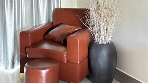 Un sofá de cuero marrón, un otoman redondo y una alta jarra con ramas, todo frente a una pared.