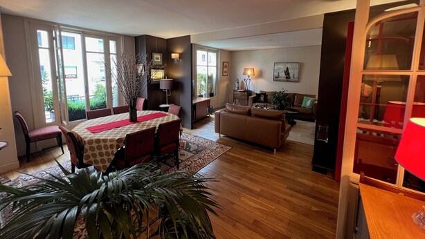 A spacious living area with a dining table, a sofa, and a coffee table. There are large windows providing natural light and a view of the outdoors. - Luxury apartment next to the Trocadéro (Paris)