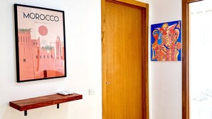 Un couloir avec une porte en bois, un tableau encadré représentant le Maroc, une petite étagère en bois et une peinture abstraite aux couleurs vives.