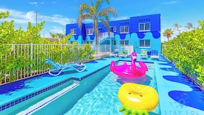 A swimming pool with inflatable toys, a white fence, and a blue building with palm trees. - 2BR Hollywood Broadwalk Haven | Free Parking (Hollywood)