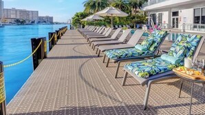 A deck with lounge chairs and umbrellas, a poolside area, and a view of the water and buildings. - Near Beach|Modern Apartment: Amazing Pool (Hallandale Beach)