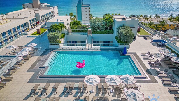 Une piscine d’un complexe hôtelier agrémentée d’un flotteur en forme de flamant rose, entourée de chaises longues et de parasols, avec vue sur une plage et l’océan au coucher du soleil.