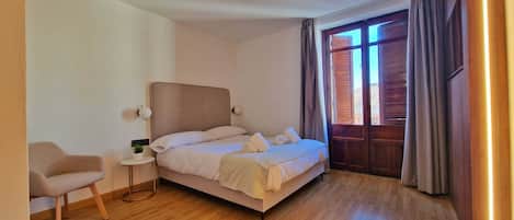 Une chambre d’hôtel moderne avec un grand lit, une petite table de chevet, une chaise et une vue sur l’extérieur par une fenêtre munie de rideaux.