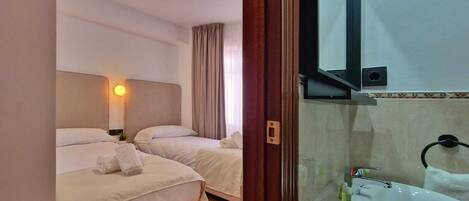 A hotel room with two beds, a bedside lamp, a window with curtains, a sink, and a mirror.