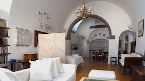 A spacious living room with a white sofa, a glass coffee table with books, and a dining area with a table and chairs. - The Palm Rock Finikia Santorini (Santorini)
