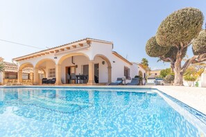 A poolside view of a building with a tiled pool, outdoor seating, and a large tree.