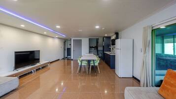 Una sala moderna con una televisión de pantalla plana, un sofá, una mesa de comedor con sillas, una cocina con refrigerador y un ventanal con cortinas.