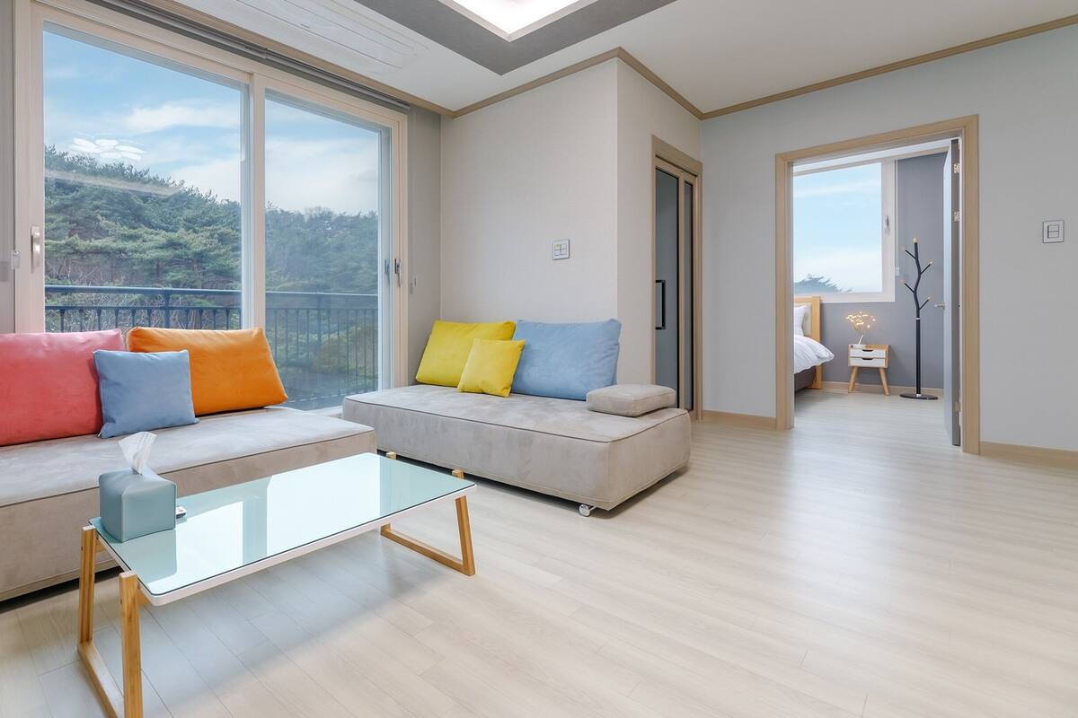 A modern living room with a glass coffee table, two sofas with colorful cushions, and large windows offering a view of greenery.