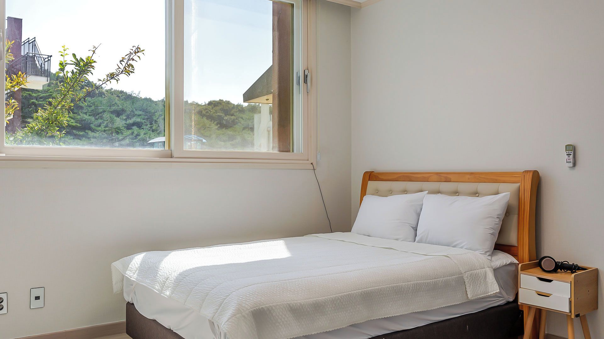 A bedroom with a bed, a nightstand, a window, and a view of greenery outside.