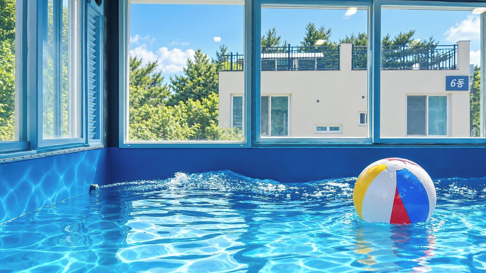 Una piscina interior con agua azul cristalina, una pelota de playa y vistas a un edificio con balcón a través de amplias ventanas.