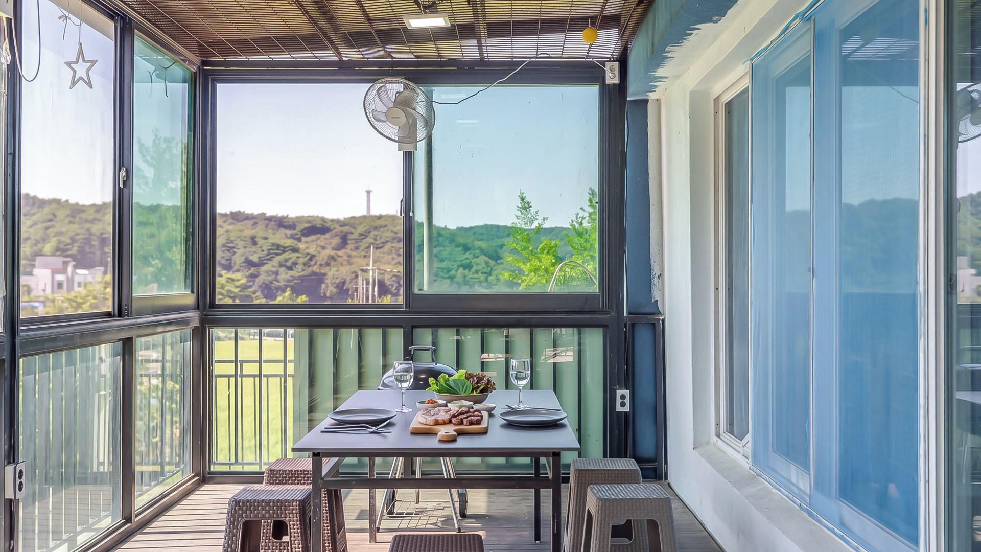 A covered outdoor dining area with a table set for two, a fan, and a view of a green landscape.