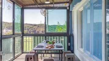 Un espacio exterior cubierto para comer con una mesa lista para dos personas, un ventilador y vistas a un paisaje verde.