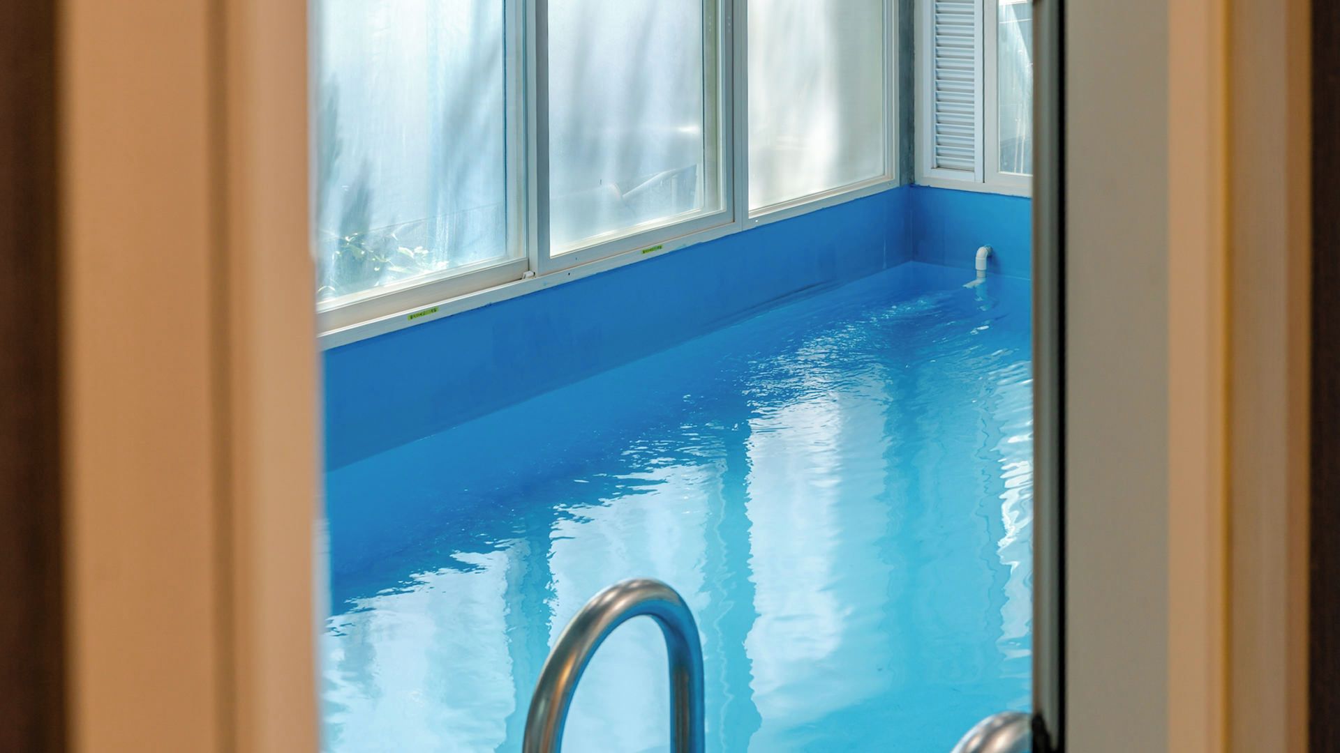 Una piscina interior con agua azul cristalina, una escalera metálica y una ventana con cortinas blancas.