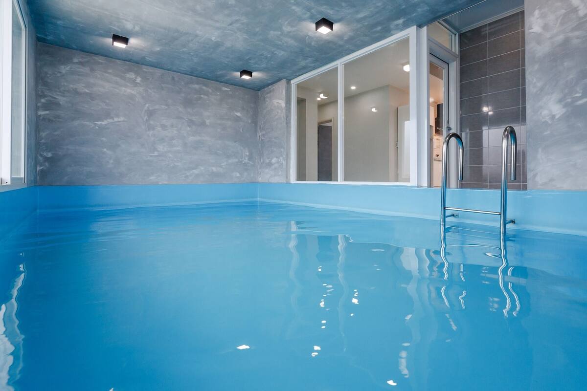 An indoor swimming pool with clear blue water, a ladder, and a modern design with grey walls and ceiling lights.