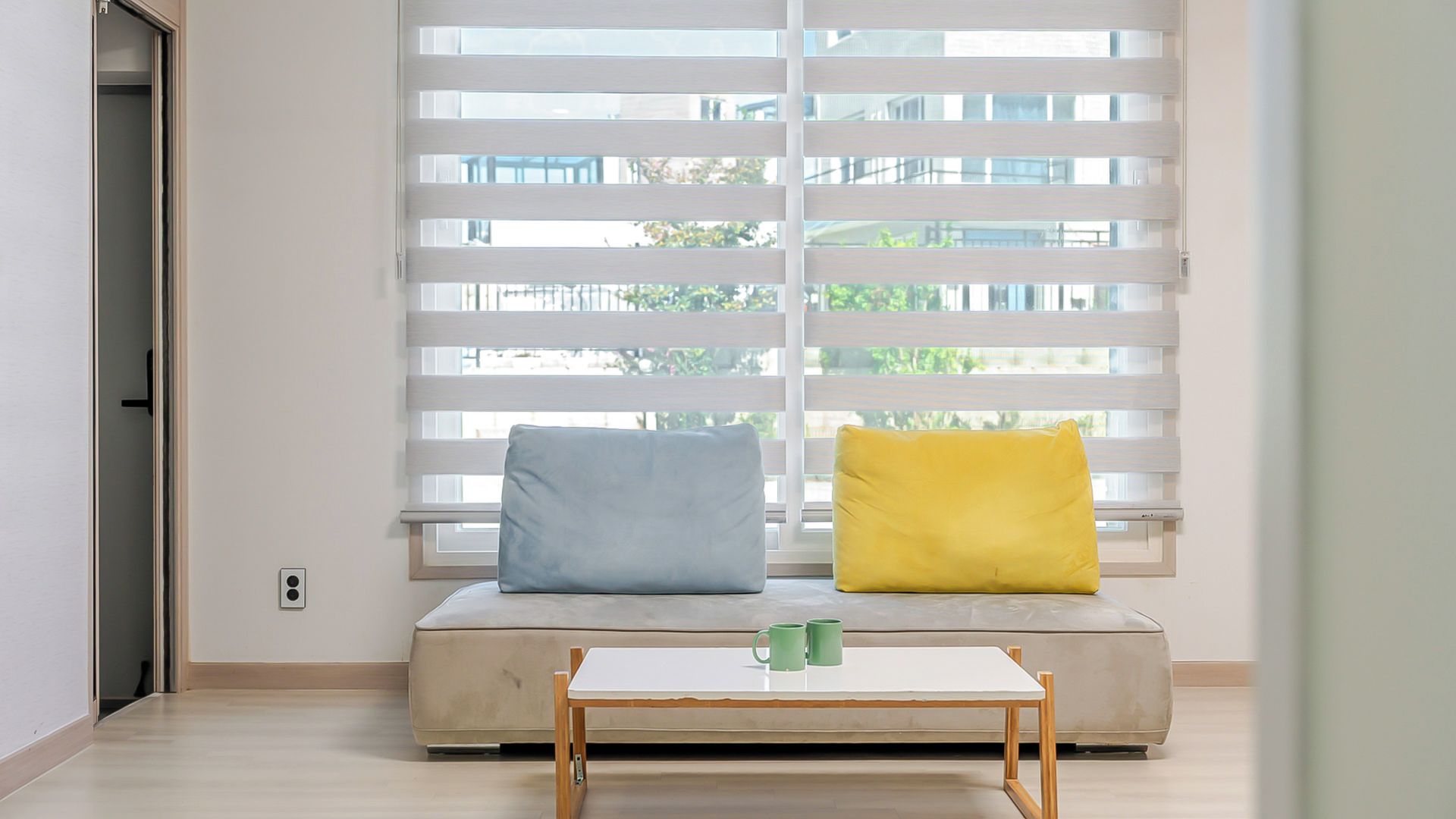A room with a sofa against a window with blinds, a small table with two mugs, and a door to the left.