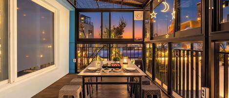 Un área de comedor al aire libre con una mesa dispuesta para dos personas, rodeada de sillas de mimbre, con vista a un atardecer.
