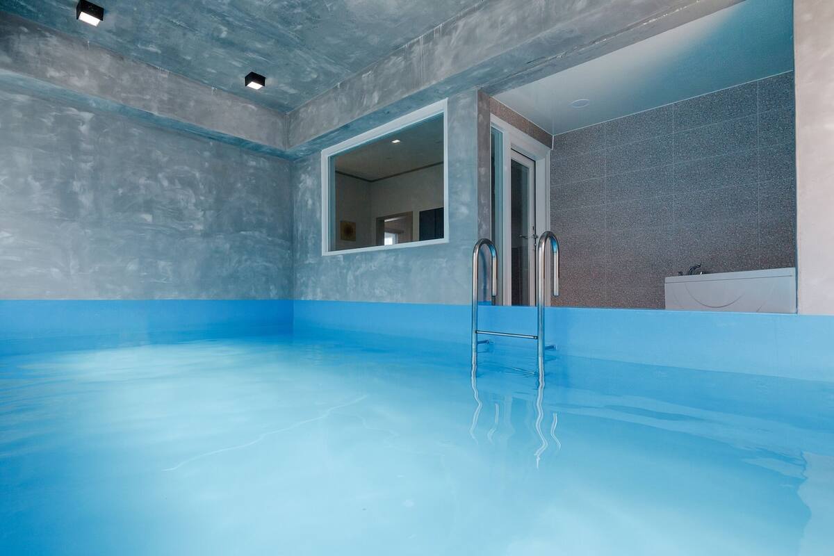 An indoor swimming pool with a blue floor, a ladder, and a bathroom with a sink and mirror.