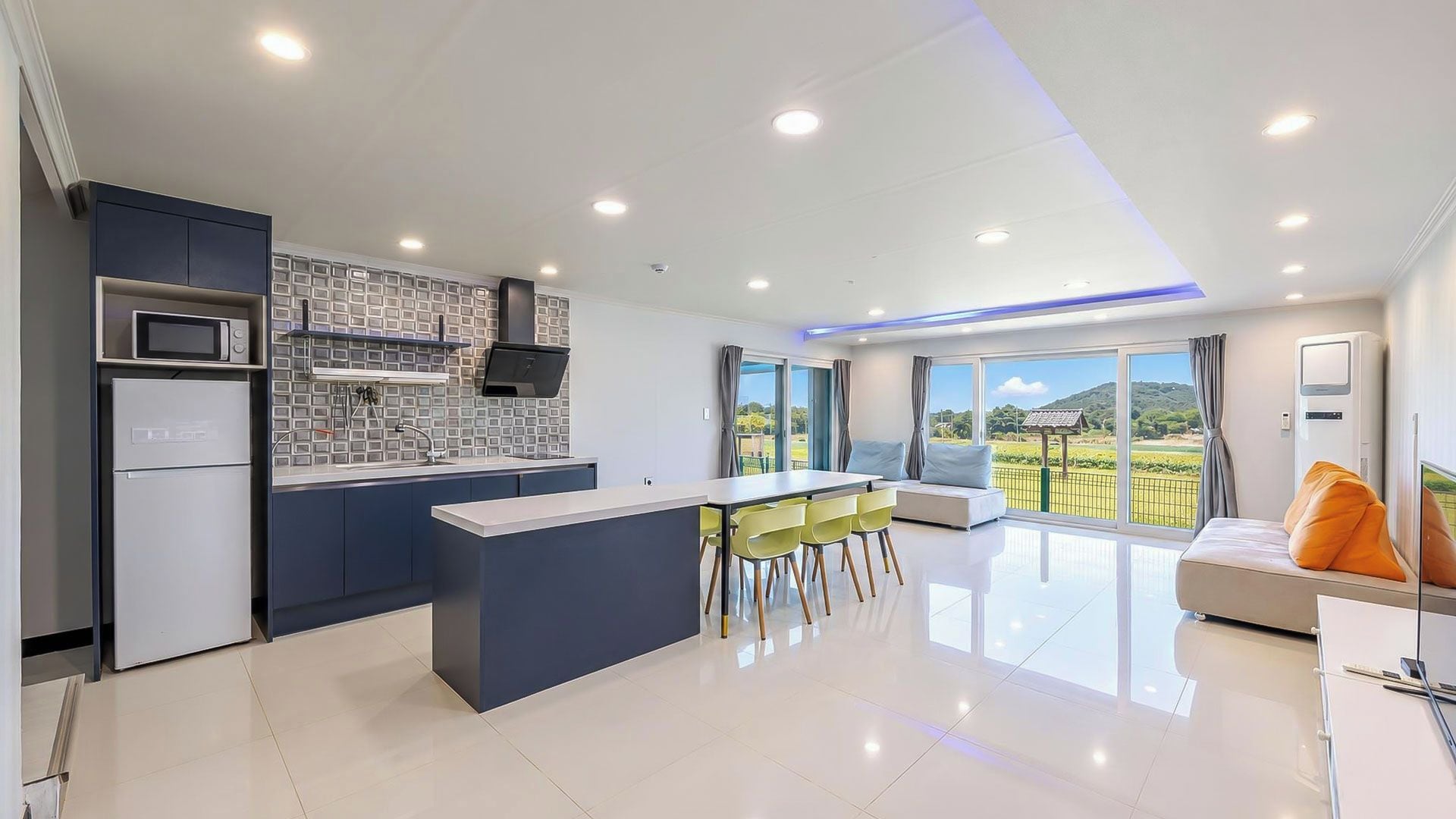 A modern kitchen with a central island, a dining area with a sofa and chairs, and a large window offering a view of the outdoors.