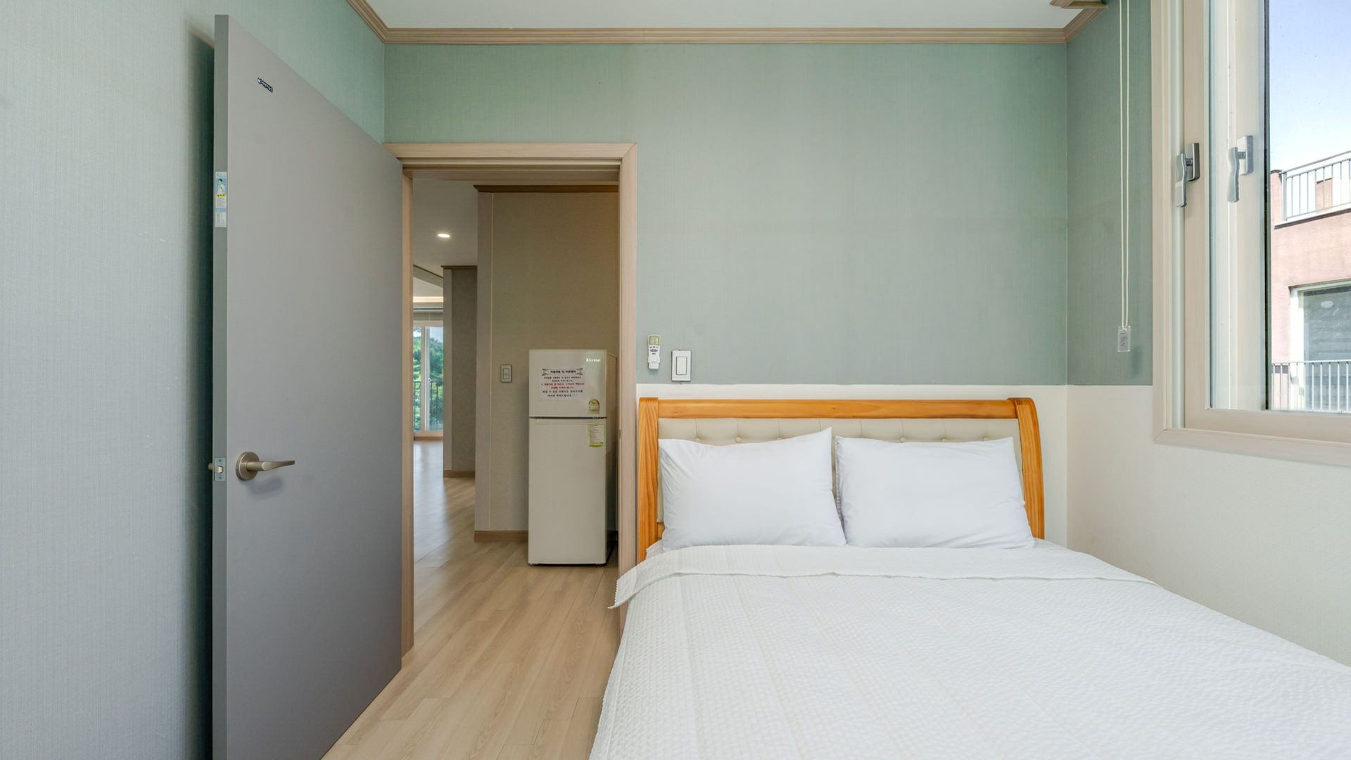 A bedroom with a bed, a door, a refrigerator, and a window.
