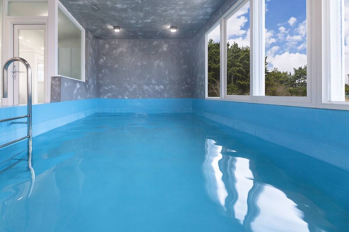 A clear indoor swimming pool with a blue interior, a ladder, and a view of trees through large windows.