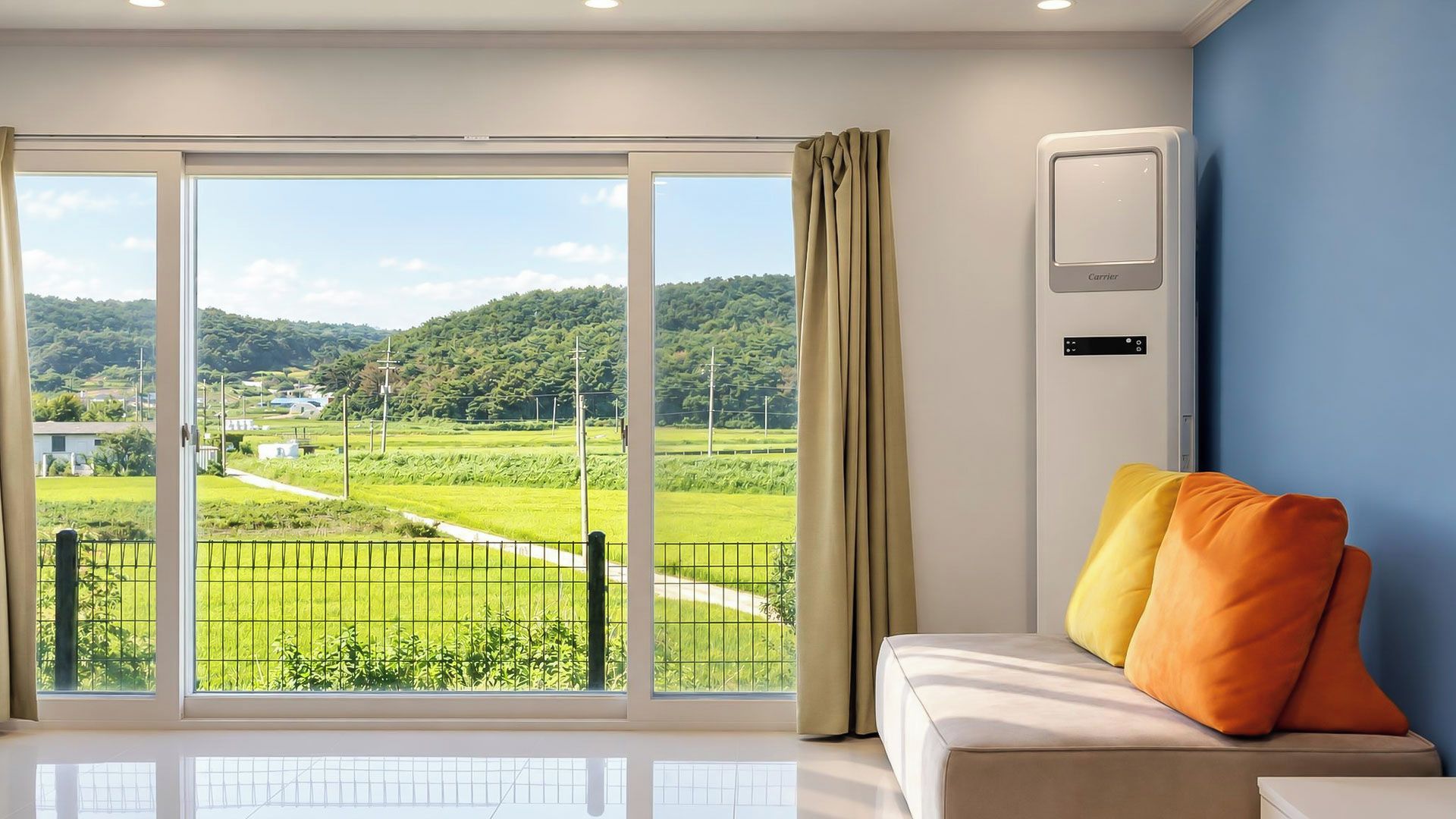Ein Zimmer mit einer großen Glasschiebetür, die einen Blick auf eine grüne Landschaft freigibt, ein weißes Sofa mit orangefarbenen Kissen und eine Wand-Klimaanlage.