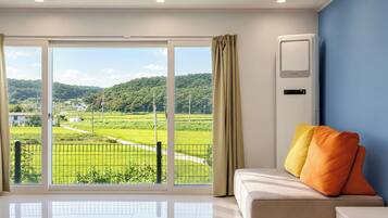 Ein Zimmer mit einer großen Glasschiebetür, die einen Blick auf eine grüne Landschaft freigibt, ein weißes Sofa mit orangefarbenen Kissen und eine Wand-Klimaanlage.